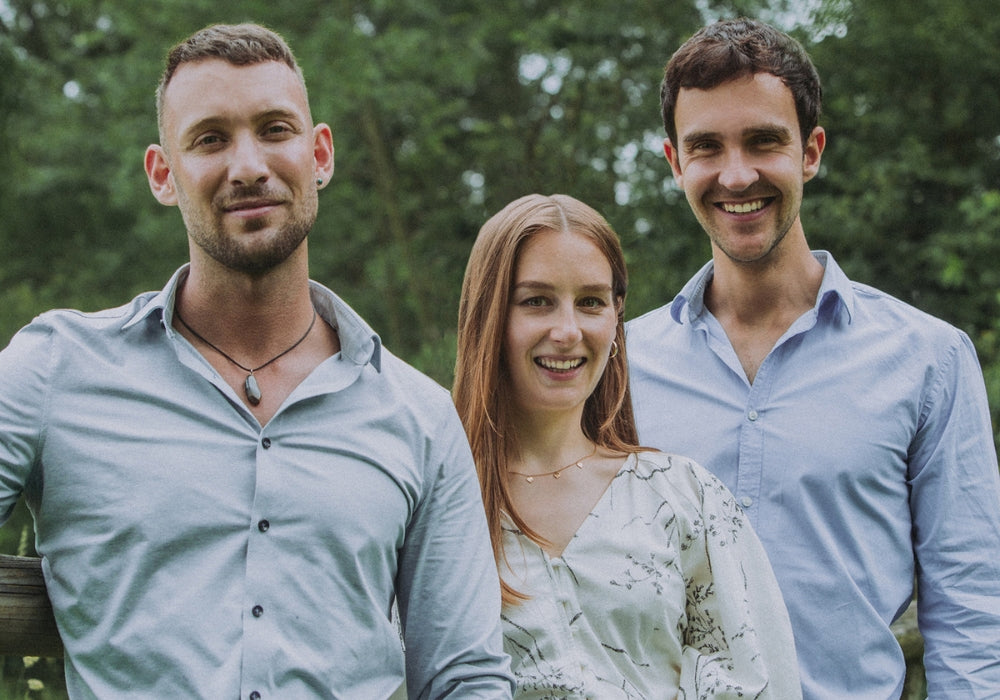 Drei lächelnde Personen im Teamfoto, draußen vor grünem Hintergrund aufgenommen