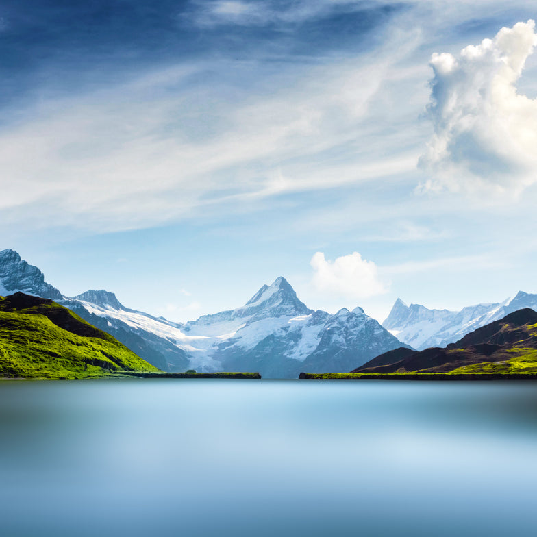Schweizer Alpenpanorama mit schneebedeckten Gipfeln, grünen Hängen und klarem Bergsee unter blauem Himmel
