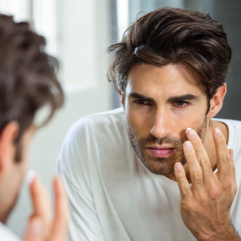 Mann trägt Gesichtscreme im Spiegel auf und überprüft dabei seine Haut im Bad